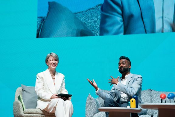 Rama being interviewed on stage by a female interviewer in a white suit.  Both are smiling into the camera.