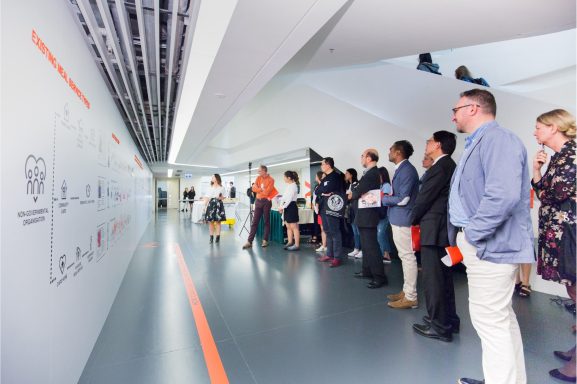 20-30 people gathered around a designer who is speaking about an idea that is depicted on a 10m long wall.